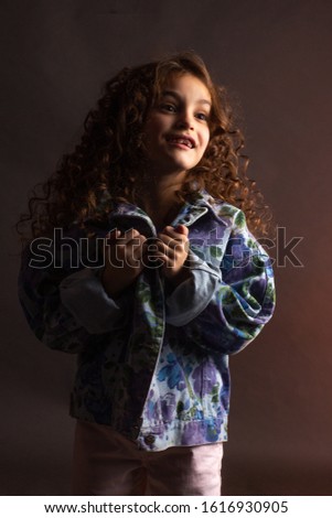Studio portrait of a little girl with long curly hair, in a colorful jacket and light shorts.