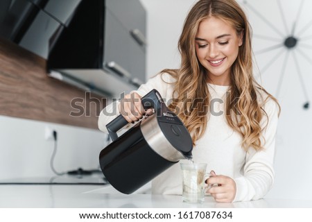 Photo of beautiful happy woman with blonde hair smiling and making tea while standing at cozy kitchen