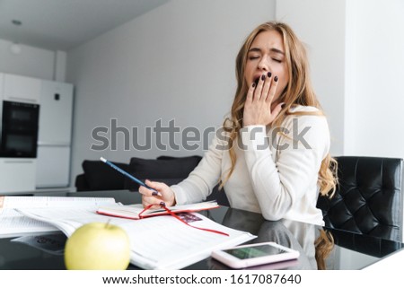 Photo of young sleepy woman yawning and making notes in exercise book while sitting at table