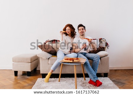 Blissful couple eating pizza in living room. Ginger girl and her boyfriend posing in cozy flat.