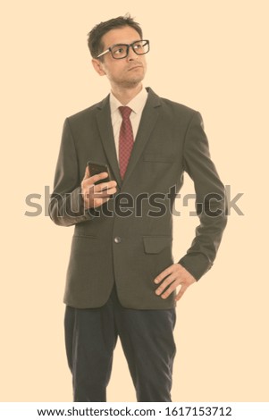 Studio shot of young businessman standing and thinking while holding mobile phone