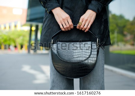 Detail of young fashionable woman wearing black blouse and grey jeans She is holding stylish crocodile print handbag in hands. Street style.