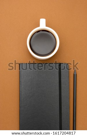 Black notebook, pencil, cup of coffee on the brown background, table top, working space with copy space for text.