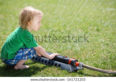 Little curly boy plays with sprinkler and looks at jets in garden at sunny summer day.