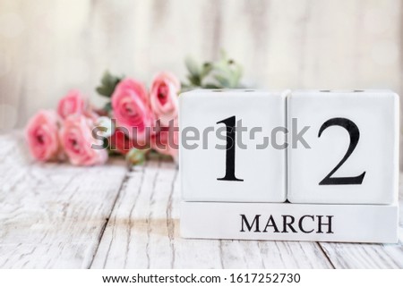 White wood calendar blocks with the date March 12 th. Selective focus with pink ranunculus in the background over a wooden table.