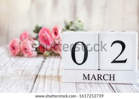 White wood calendar blocks with the date March 02. Selective focus with pink ranunculus in the background over a wooden table.