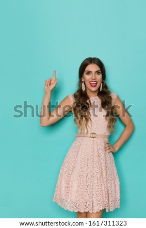 Happy beautiful young woman in elegant beige lace mini dress is pointing up, looking at camera and talking. Front view. Three quarter length studio shot on turquoise background.
