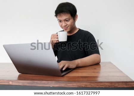 Young Asian man feeling happy and smile what he see in laptop with drink coffee in white glass. Indonesia Man wear black shirt Isolated grey background.