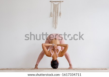 Young unidentified slim woman brunette yoga lover in pink costume doing prasarita padottanasana while standing on rug on floor on white wall. Yoga instructor does an advanced asana. Advertising space