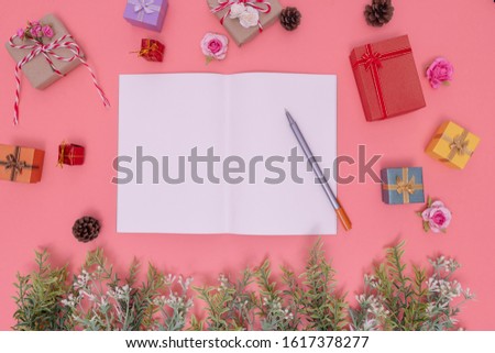 various gift boxes and artificial green plants put around the picture and white paper and pen place in the center. those are placed on a pink background