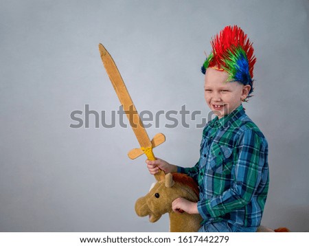 happy boy in plaid shirt is rolling on rocking horse toy. baby with wonderful punk-style colored hairdo. kid is armed with wooden sword and brandishes it. Dreams about travel and heroic exploits
