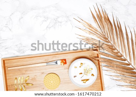 Wellness spa concept with body cream and golden pieces, washcloth, massage brush, massage oil jar, golden palm leaf on wooden and marble backdrop. Advertising template for salon, skin care, wellbeing