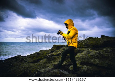 Outdoor photographer with contrast yellow jacket looking photos that he shot over rocks among waves gloomy sky with dramatic clouds