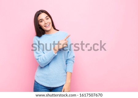 young pretty woman looking excited and surprised pointing to the side and upwards to copy space against pink wall