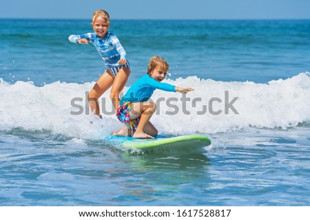 Happy baby boy and girl - young surfers ride with fun on one surfboard. Active family lifestyle, kids outdoor water sport lessons, swimming activity in surf camp. Sea beach summer holiday with child.