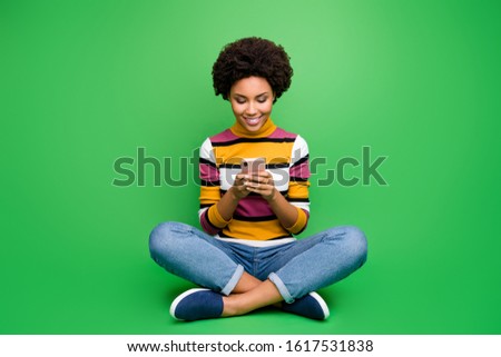 Full length photo of positive afro american girl blogger sit legs crossed folded use cellphone post comment share repost wear bright shine outfit isolated over green color background