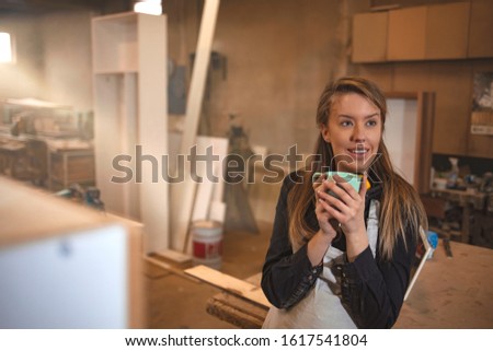 Mid Adult Female Carpenter Working In Her Workshop. Standing by the desk and drinking coffee. Woman carpenter making a coffee break, drinking coffee. Woman carpenter doing regular day office work