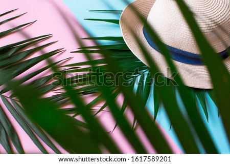 a beach hat lies on a green tropical leaf on a pastel pink-blue background, the effect of peeping through palm leaves is created