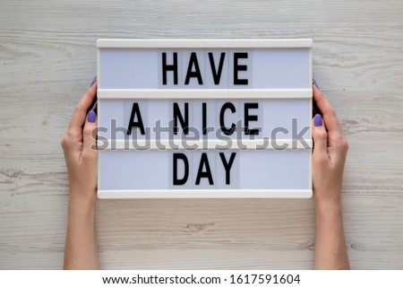 Female hands hold a lightbox with 'Have a nice day' words on a white wooden background, top view. Overhead, from above, flat lay. 