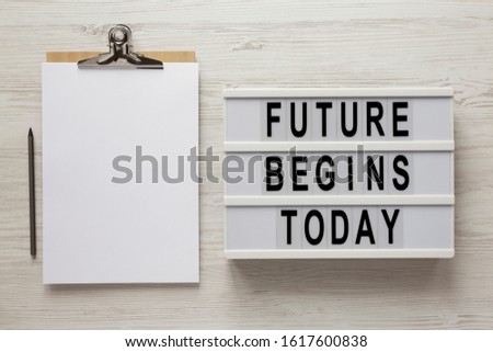 'Future begins today' words on a lightbox, clipboard with blank sheet of paper on a white wooden surface, top view. Overhead, from above, flat lay. 