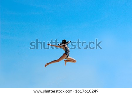 Happy young girl jumping up to the sky. People, happiness, travel concept. Attractive teenage girl wearing a big black sun hat and swimsuit  jumping over blue sky background.
