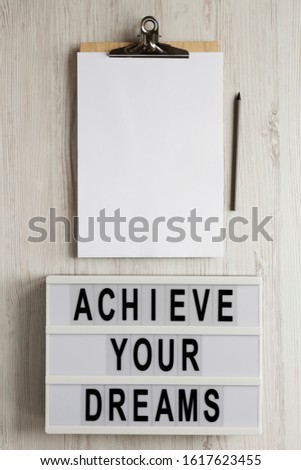 'Achieve your dreams' words on a lightbox, clipboard with blank sheet of paper on a white wooden background, top view. Overhead, from above, flat lay. Copy space.