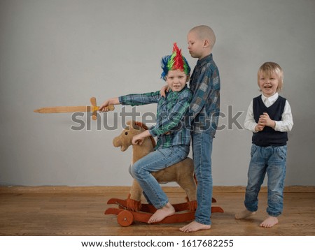 Two younger brothers pushes boy in plaid shirt, who is rolling on rocking horse toy. baby with beautiful punk haircut. kid is armed with wooden sword and brandishes it. Dreams of travel, heroic deeds