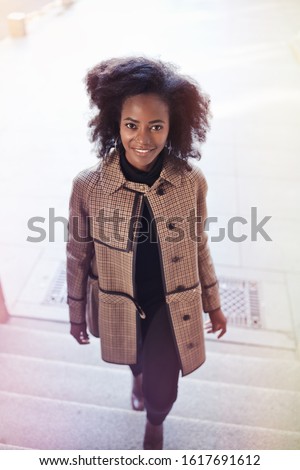 Beautiful african american young woman with afro and large hoop earrings in a stylish coat, smiling. Urban street portrait. Toned picture.