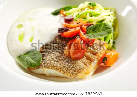 Cod fillet in cream sauce with vegetable spaghetti, fresh greens on white restaurant plate isolated. Macro shot of breaded white fish meat with cherry tomatoes, olives and spinach closeup