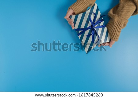 both hand holding a blue and white striped gift box tied with ribbon on the top left corner of a picture on blue background