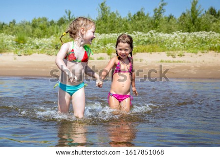 Little girl friends run along the river and spray water on a sunny summer day.
