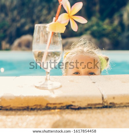 Cute little blonde girl swimming. Big glass with water, straw and frangipani stay on the pool edge. Little lady peeps from pool and look to the camera. Sunbathing and leisure on sunny summer day.