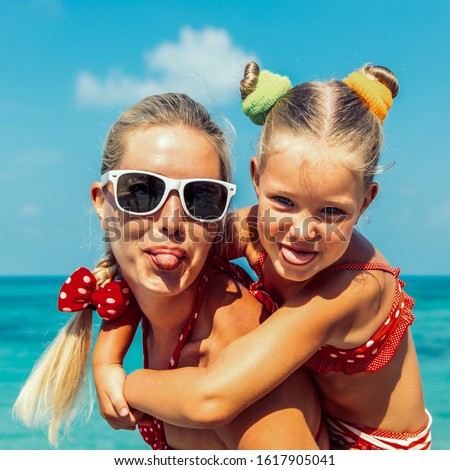 Beautiful mom in sunglasses with cute little daughter on her back. Two in red bikini. Woman and little lady show tongue, smile and looking to camera. Happy family on the sea shore. Happy mothers day.