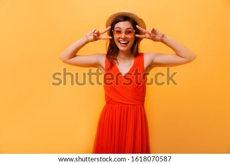 Indoor portrait of European girl isolated on yellow background in casual clothes with optimistic smile, showing victory sign with both hands, looking friendly and willing to welcome and communicate.