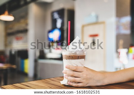 cold drink in transparent glass with whipped cream on top in girl's hand on blurred restaurant background