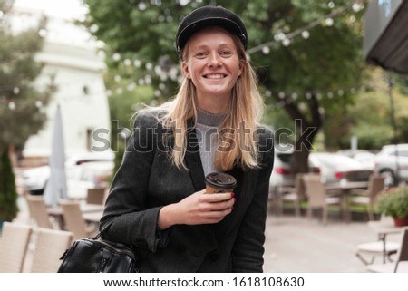 Charming young happy blonde long haired lady in black hat and elegant blazer posing over urban background with paper cup in raised hand, smiling widely while looking at camera