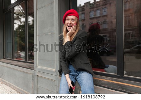 Amazed attractive young blonde female with red manicure looking surprisedly in front of herself and holding palm on her cheek, posing outdoor in trendy clothes