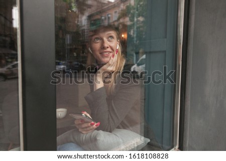 Attractive young blue-eyed blonde lady in elegant clothes looking dreamily out window while sitting over windowsill, wearing headphones and holding mobile phone