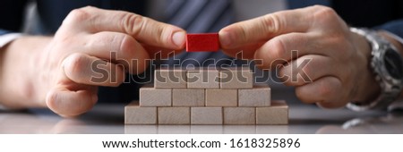 Close-up view of presentable man putting unique red block on staircase of other identical bricks. Pyramid of wooden cubes on desktop. Authority and uniqueness concept Royalty-Free Stock Photo #1618325896