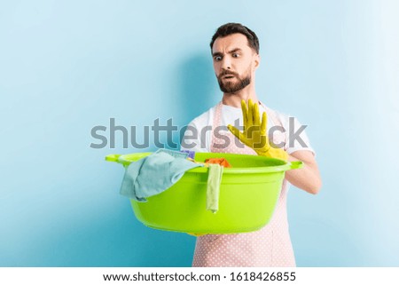 shocked man holding plastic wash bowl with dirty laundry and showing stop gesture on blue