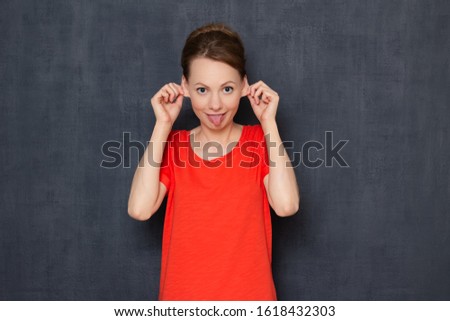 Studio portrait of funny happy young blond woman wearing orange T-shirt, touching ears with hands and sticking out her tongue, smiling, fooling around and having fun, standing over gray background