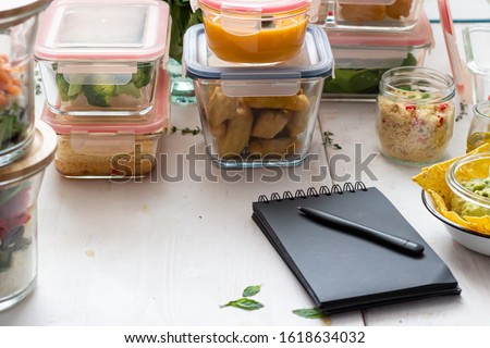 Meal prep scene with piles of glass jars full of homemade meal. A black notepad with a pen on it is in the front. Royalty-Free Stock Photo #1618634032