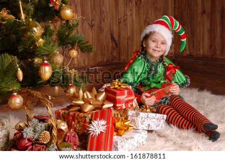 Girl in suit of Christmas elf with a gift near Xmas fir-tree