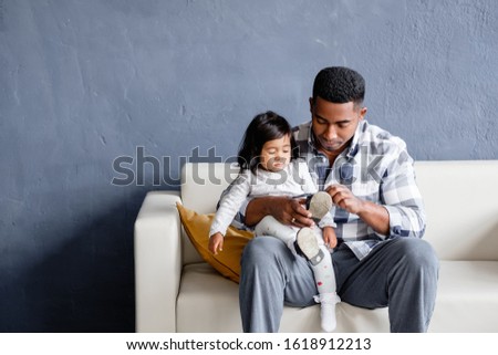 Caring mixed race father holds his pretty little daughter in his arms and puts on her shoe while sitting in a cozy sofa in the living room. Concept of love between father and daughter. Copyspace
