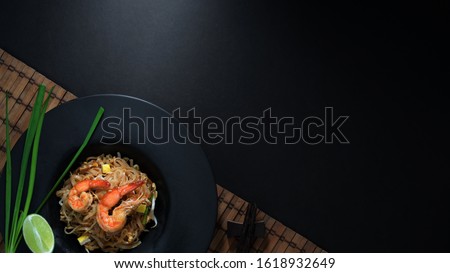 Overhead shot of Pad Thai, stir fly of Thai noodle with shrimp and egg in black ceramic plate on bamboo placemat with copy space on black table