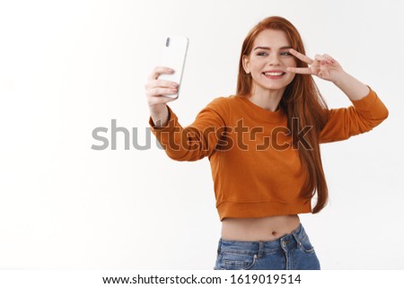 Carefree pretty millennial hipster girl with red long hair taking selfie using white smartphone, make peace sign and smiling, photographing, capture happy moment, white background