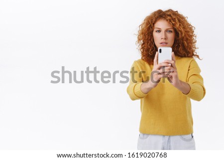 Focused, serious-looking redhead curly female in yellow sweater squinting, look determined at smartphone display as record video or trying take good shot, photographing something, white background