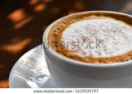 Cover the white coffee cup and foam latte on a black wooden table near the window, with light shades on the coffee shop table.