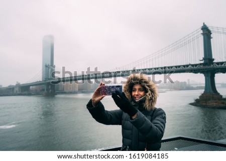 Tourist woman with warm clothes taking a selfie with the Manhattan Bridge on the background in DUMBO neighborhood while sightseeing new york during winter season. teal and orange style