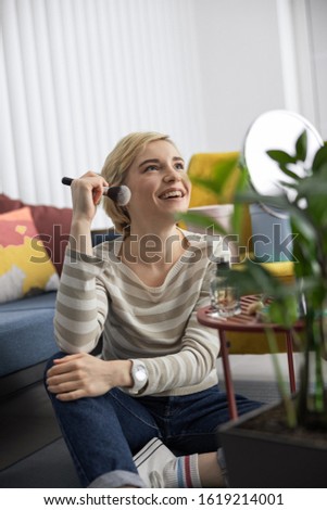 Smiling attractive woman sitting on floor while doing makeup at home stock photo
